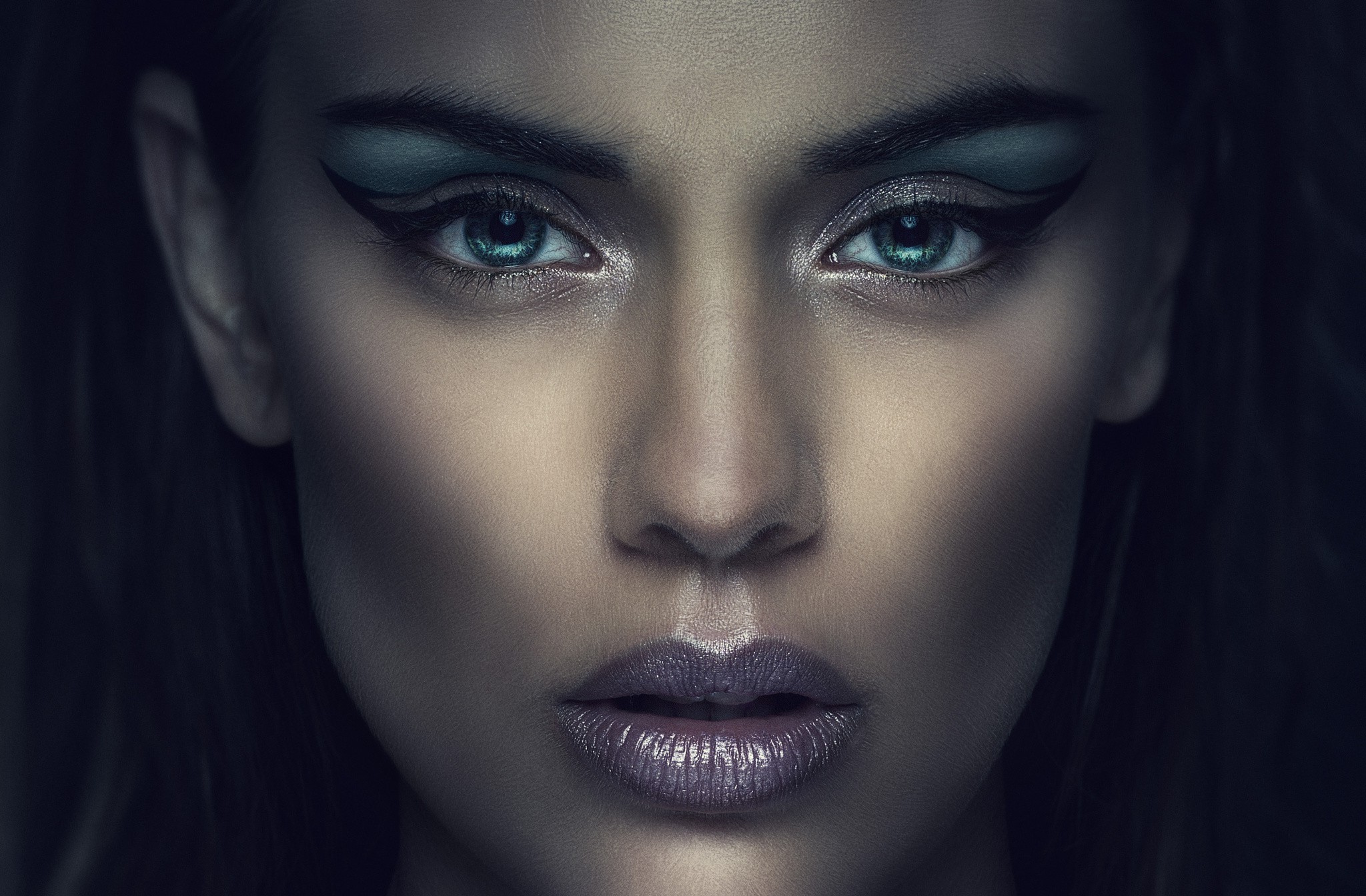  A closeup of a woman's face with clear and glowing skin, dark hair, and dark eyeshadow.